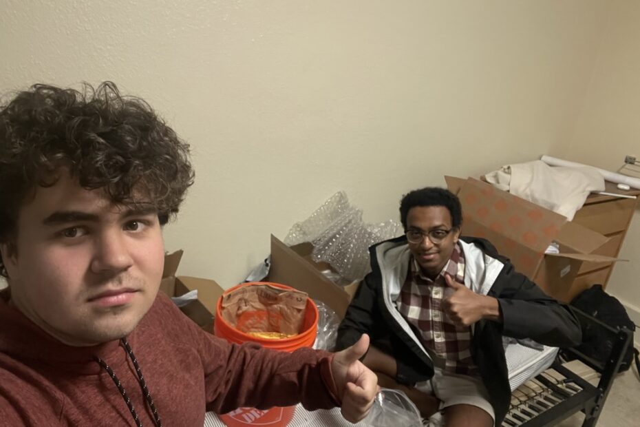 two students give thumbs up signs, they sit among several open boxes and packaging material.