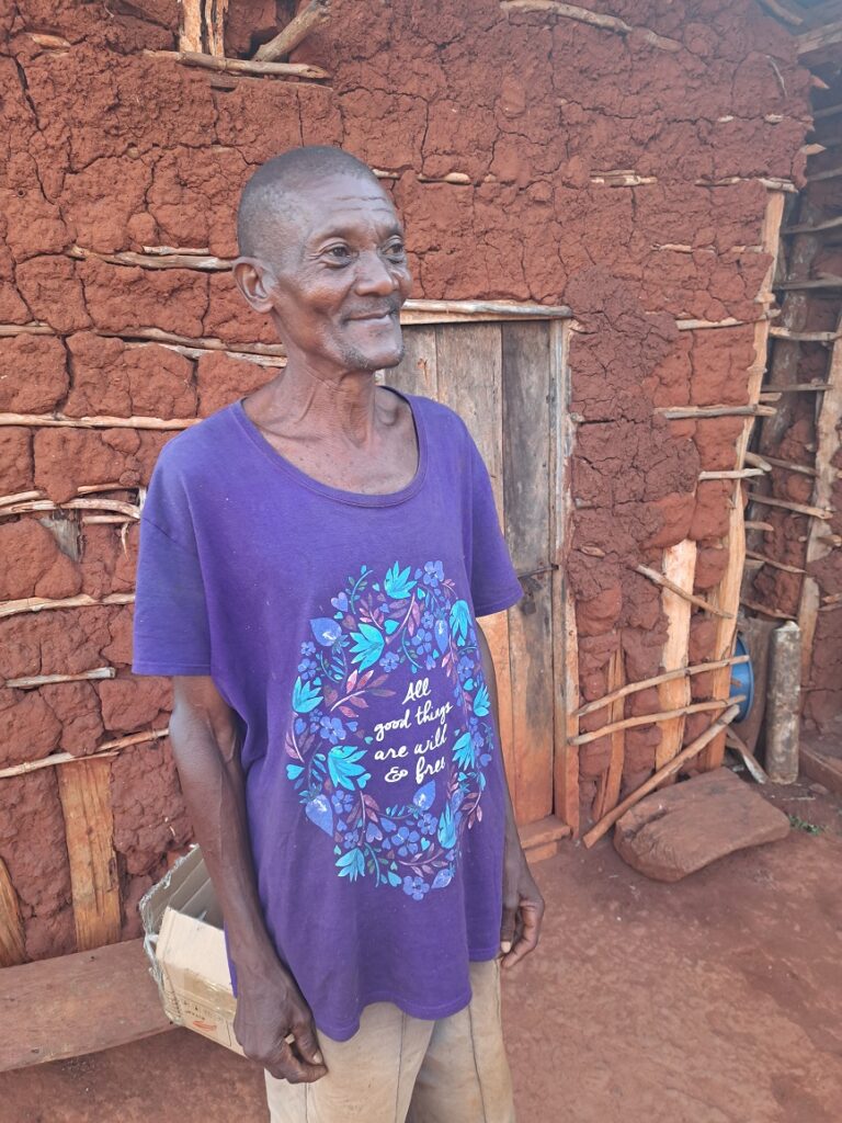 A smiling villager in a purple shirt with an English saying on it, in front of a mud house with a wooden door.