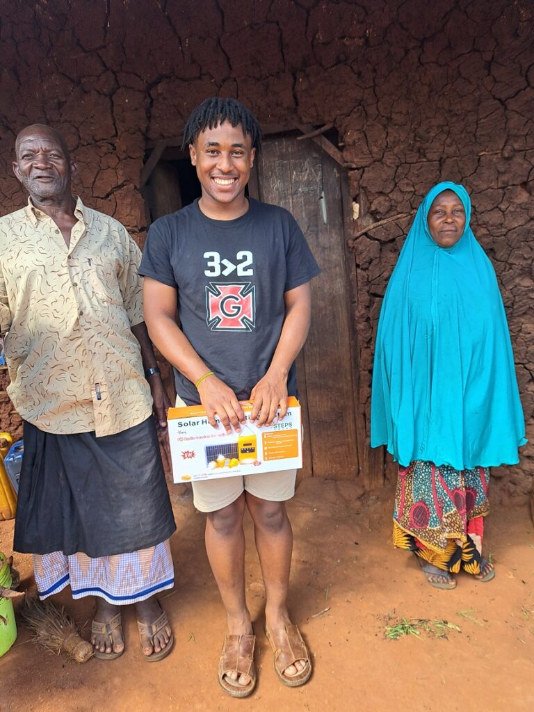 Villagers, in front of a mud house with a wooden door, with a box of solar light equipment.